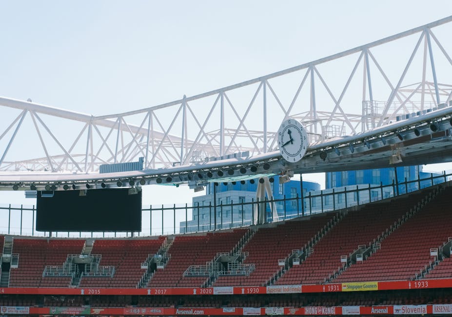 Interior view of a sports stadium during daytime showing rows of red seating with some empty seats, a large digital scoreboard, and a prominent white clock mounted on a structural beam of the roof. The roof is supported by a white truss framework that spans across the open-air arena, allowing natural light to illuminate the scene. In the foreground, several cardboard boxes and plastic wrapping materials are arranged on the pavement near the entrance, suggesting a packing or loading process associated with home relocation or moving services. Some boxes are stacked, while others are being carried or positioned with a trolley. The scene hints at furniture transport or packing activities involved in house removals, with the stadium environment providing a neutral background. Removals Highbury occasionally operate in large-scale moving logistics, as reflected in the organized packing materials and loading area visible in the image.