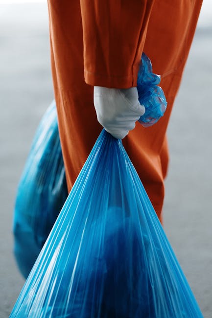 A person wearing orange pants and white sneakers is standing on a gray carpeted floor, holding two large blue plastic garbage bags filled with household waste or packing materials. One bag is in each