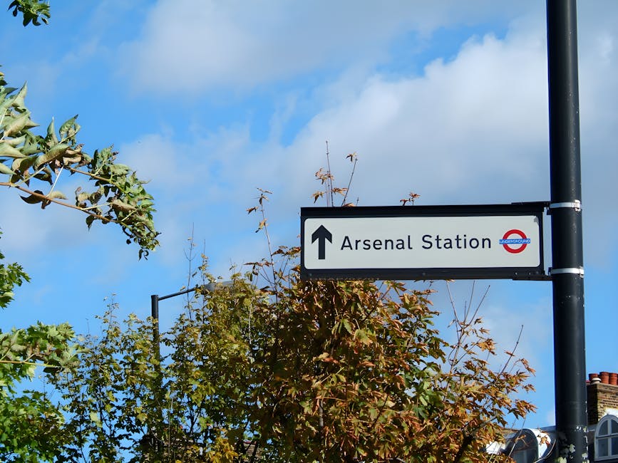 A rectangular directional street sign attached to a black metal pole indicates the way to Arsenal Station. The sign features a white background with black text and a black upward arrow, along with the