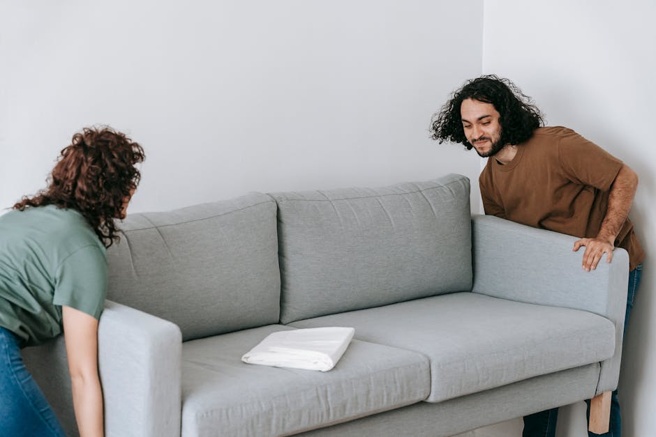 Two individuals are engaged in a home relocation process inside a modern living room, preparing to transport a light gray fabric sofa with wooden legs. The woman with curly brown hair, wearing a green T-shirt and jeans, is leaning forward, adjusting or securing the sofa, while the man with curly black hair, dressed in a brown T-shirt and jeans, is positioned behind the sofa, holding onto its armrest. On the sofa, there is a folded white blanket or protective padding, likely used to cushion furniture during packing and moving. The room features plain white walls and minimal decor, with natural lighting illuminating the scene, emphasizing the careful handling of the furniture as part of the packing and moving activities. This image reflects the logistical aspect of furniture transport involved in house removals, captured during the loading process, demonstrating the work of a professional removal service such as Removals Highbury.