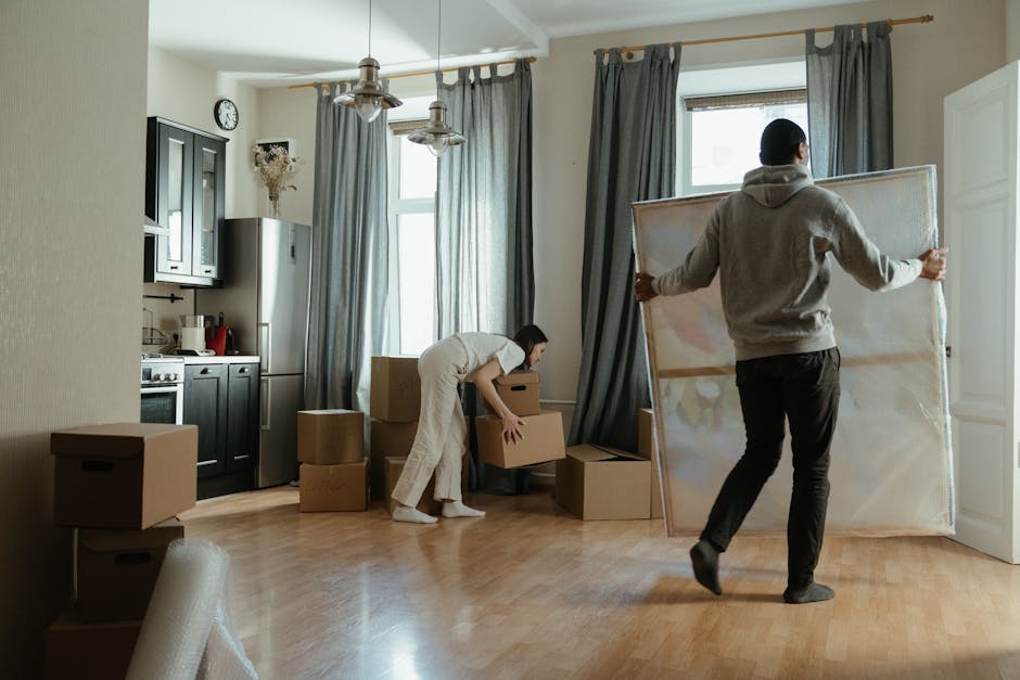 Two movers from Removals Highbury are inside a well-lit, spacious apartment preparing for a home relocation. One individual, wearing a grey hoodie and black trousers, is carrying a large, framed mirror by holding its edges, positioning it to be moved through an open door. The other worker, dressed in casual light-coloured clothing, is crouched down, lifting a medium-sized cardboard box from a pile of packed boxes and packing materials on the wooden floor. Several closed cardboard boxes of varying sizes are stacked against the wall near the kitchen area, which features dark cabinetry, a stainless-steel refrigerator, and small kitchen appliances on the countertop. The room has two large windows with grey curtains, allowing natural light to fill the space. The scene involves packing and lifting activities as part of furniture transport and packing and moving processes supported by the moving company during a house or apartment relocation.