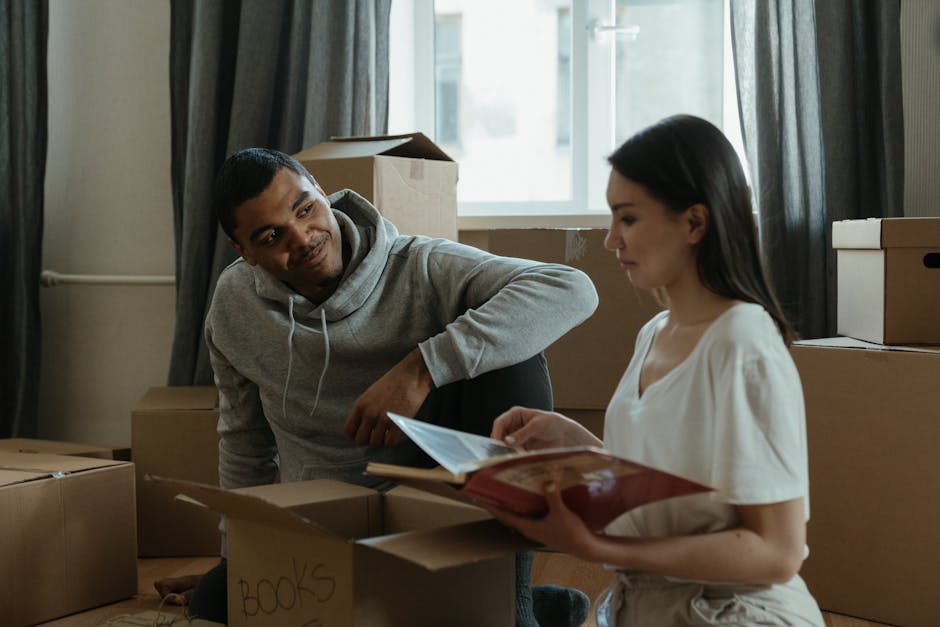Two individuals are engaged in a home relocation process inside a modern living room, preparing to transport a light gray fabric sofa with wooden legs. The woman with curly brown hair, wearing a green T-shirt and jeans, is leaning forward, adjusting or securing the sofa, while the man with curly black hair, dressed in a brown T-shirt and jeans, is positioned behind the sofa, holding onto its armrest. On the sofa, there is a folded white blanket or protective padding, likely used to cushion furniture during packing and moving. The room features plain white walls and minimal decor, with natural lighting illuminating the scene, emphasizing the careful handling of the furniture as part of the packing and moving activities. This image reflects the logistical aspect of furniture transport involved in house removals, captured during the loading process, demonstrating the work of a professional removal service such as Removals Highbury.