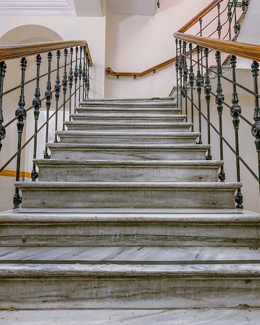 A set of indoor marble stairs with polished, slightly worn steps leading upward, flanked by black and brass railings with decorative balusters. The stairwell has white walls, with a wooden handrail running along both sides. The space is well-lit, possibly by natural light, and the image captures the stairs from the bottom looking upward. In the context of house removals, the image exemplifies navigating narrow staircases during a home relocation or furniture transport, part of the loading process managed by Removals Highbury, a professional moving service specializing in house removals and packing logistics.