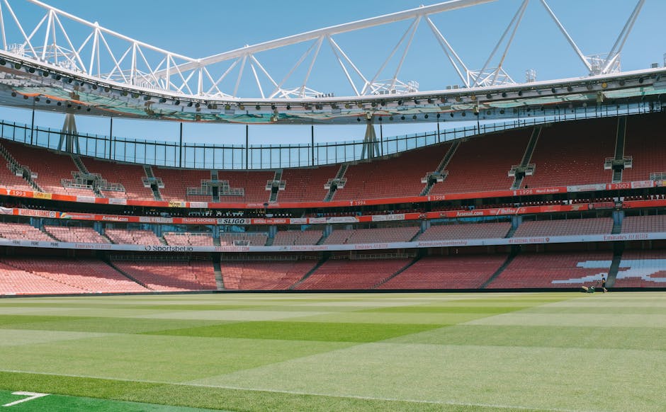 The image shows a large, modern football stadium with tiered seating sections filled with red and pink seats, surrounding a well-maintained grass pitch with visible marking lines. The interior is open to a blue sky, and a prominent white steel roof structure spans across the top, supported by cables and beams. The scene appears well-lit by natural daylight, and there are no spectators or players present. The stadium is empty, and the view is from a perspective within the seating area, looking across the field towards the opposite stands. This environment exemplifies a professional sports venue suitable for major football matches or events. For house removals and furniture transport, understanding such venues highlights the scale and logistical considerations involved in moving bulky furniture or equipment into large-scale sporting facilities, supported by professional services like Removals Highbury, which specialise in packing, loading, and transporting large items during home relocation projects.