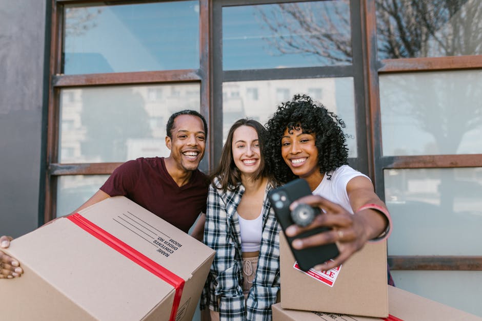 Three individuals standing outdoors in front of a building with large windows, smiling and taking a selfie together. They are dressed casually; the person on the left, a man, is wearing a maroon t-shirt and holding a cardboard box wrapped with red tape. The person in the middle, a woman, has long brown hair, and is wearing a black-and-white checkered shirt, holding another cardboard box. The person on the right, a woman with curly black hair, is wearing a white t-shirt, holding a smartphone to capture the selfie, with her arm extended. The scene is well-lit with natural daylight, and the background shows an outdoor urban environment with some trees and buildings visible through the glass windows. This image reflects the friendly aspect of home relocation and packing, as part of the packing and moving process that Removals Highbury provides, illustrating the human side of furniture transport and customer engagement prior to a professional move.