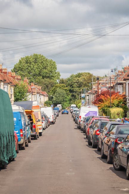 A residential street with parked cars on both sides, including a range of models such as hatchbacks and vans, some covered with protective tarpaulins. In the foreground, a Van belonging to Removals Highbury is positioned on the right side, partially visible, with a loading ramp extending from its rear. Behind the van, several cardboard moving boxes and furniture wrapped in plastic or moving blankets are being loaded or unloaded, with visible hands and lifting equipment like trolleys and straps assisting the process. The street is lined with typical British terraced houses with brick facades, some with small front gardens, and mature trees providing greenery. Overhead, power lines run parallel to the street beneath an overcast sky with grey clouds. The scene captures a moment of home relocation activity, emphasizing furniture transport and packing procedures associated with removals, aligning with peak season delays for high-volume moving schedules.