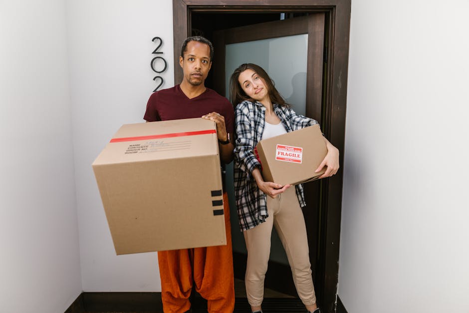 A man and a woman stand inside a residential building next to a doorway, each holding a cardboard box. The man, dressed in an orange pant and a dark red T-shirt, holds a large box with red and black markings, while the woman, wearing a plaid shirt and light-colored pants, holds a smaller, labeled fragile box. Both individuals appear to be involved in a home relocation process, with the woman leaning her head on the door frame in a relaxed manner. The scene suggests the loading or unloading phase of a furniture transport or packing and moving service, with visible interior lighting and a clean, minimalist environment. The setting aligns with professional removals activities offered by Removals Highbury, focused on house or apartment moves in the vicinity of Chapel Market to Canonbury, supporting efficient and careful furniture transport and packing logistics.